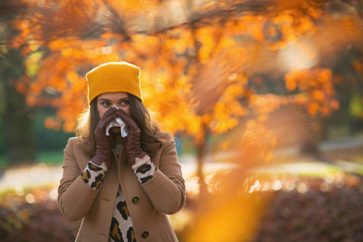 Hello autumn. modern woman in brown coat and yellow hat with napkin blowing nose outdoors in the city park in autumn.