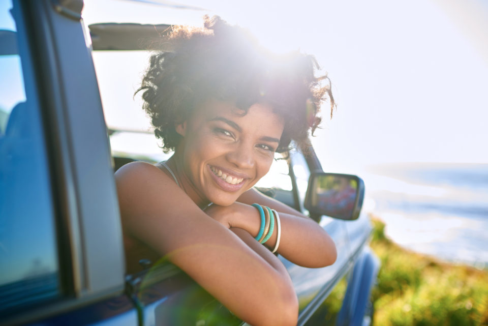 A young woman leaning out of her car while the summer sun shines behind her.