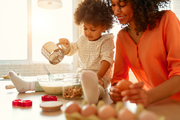 African American woman with her little daughter with curly fluffy hair having fun and cooking pastries in the kitchen. Mom and daughter cooking together. Child pouring and sifting flour into a bowl.